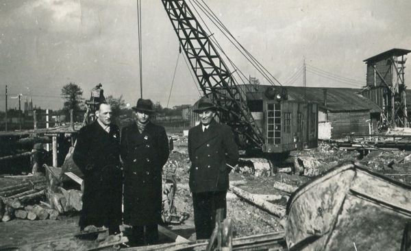 Fotografia przedstawia trzech mężczyzn na placu budowy, za nimi dźwig