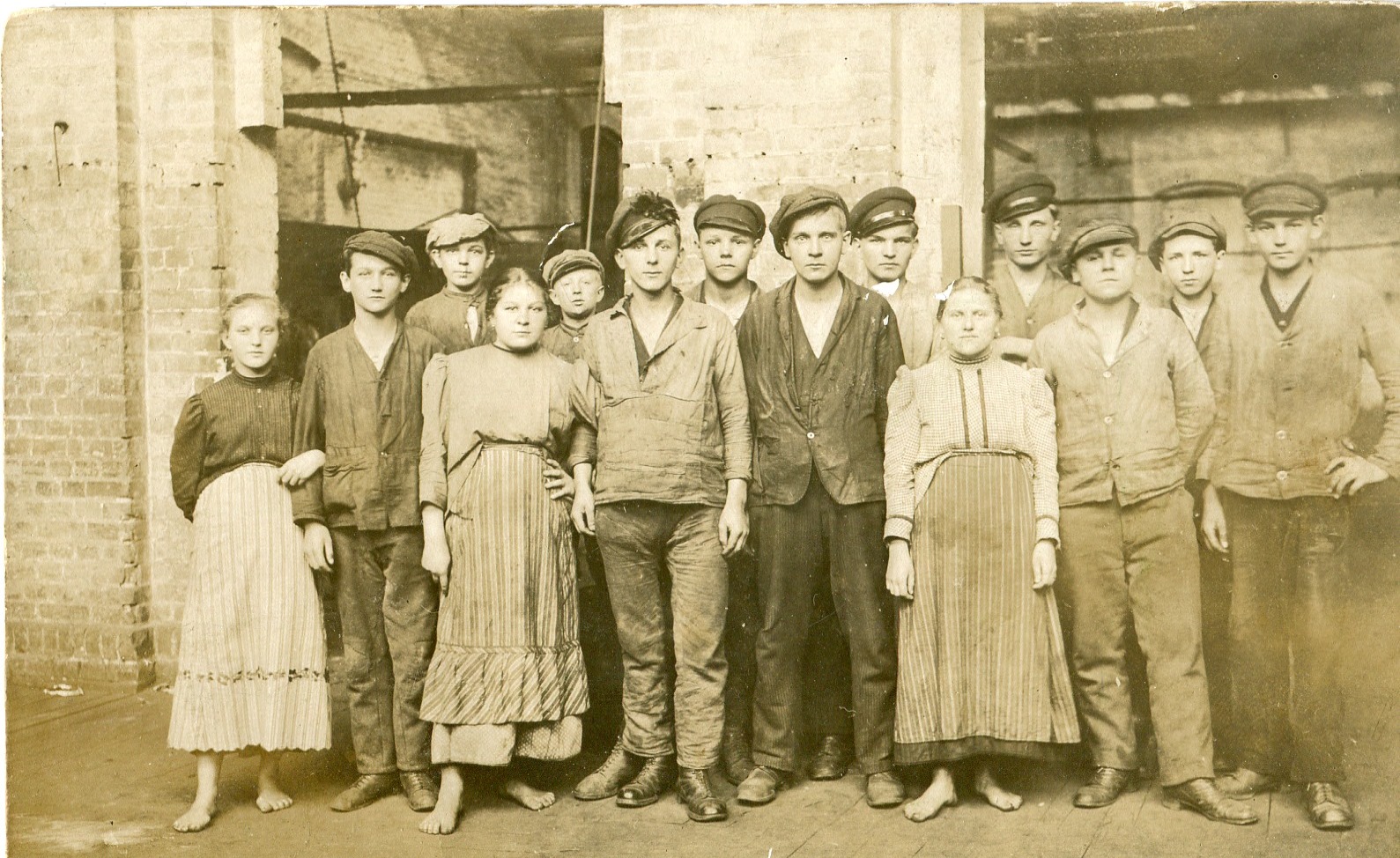 Fotografia grupy mężczyzn i kobiet w ubraniach roboczych, z pocz. XX w. na tle budynków przemysłowych
